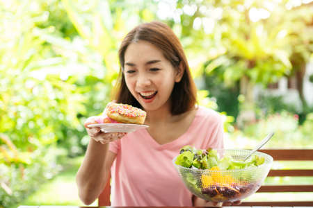 The happy beautiful Asian healthy middle aged woman sitting on the balcony beside the garden and choose between a donut and a vegetable salad. Concept of health care and nutritious foodの写真素材