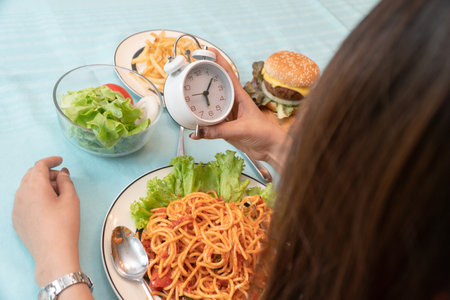 Young woman holding clock and ready to eating a hamburger, French fries, and Spaghetti for Breakfast. Concept of binge eating disorder (BED) and Relaxing with Eating junk food and unhealthy foods.の写真素材