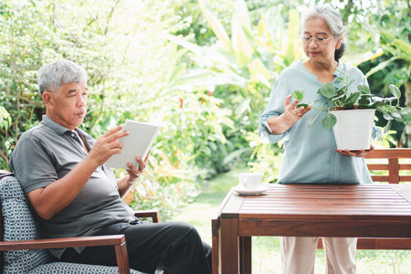 A happy and smiling Asian old elderly woman is planting for a hobby after retirement and her husband is reading a book. Concept of a happy lifestyle and good health for seniors.の写真素材