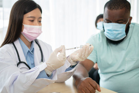 Asian doctor user syringe needle for injection vaccinate,  African American is being covid...