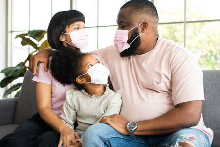 Mixed Race family sitting on the sofa and wearing medical face masks for against  world pandemic and stay quarantined together at home social distancing new normal lifestyleの写真素材