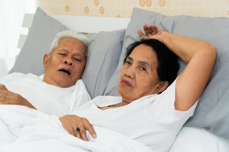 Older Asian woman laying open her eyes in bed beside her husband who snores and Makes noise, Marital problems.の写真素材
