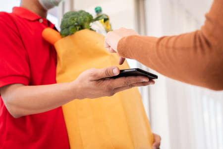 Asian delivery man holding holding a bag of fresh food for giving to customers and holding smartphone for Receive payments at home. Concept of express grocery service and new lifestyleの写真素材