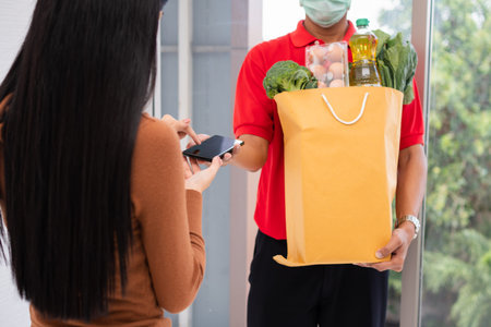 Asian delivery man holding holding a bag of fresh food for giving to customers and holding smartphone for Receive payments at home. Concept of express grocery service and new lifestyleの写真素材