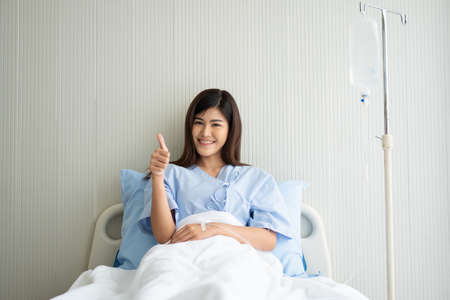 Happy Asian female patient smiling, lies on the bed, and raises thumb up to show confidence in treatment. Concept of believe in treatment And insurance coverageの写真素材