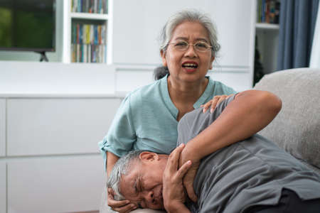 Asian old elderly man is In pain with his hands on his chest and having a heart attack in the Living room and his wife is shocked and confused, helpless. Concept of healthcare and Elderly caregiversの写真素材