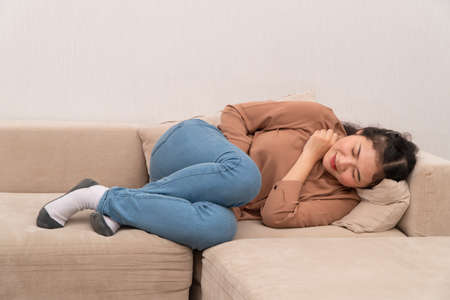 Unhappy Asian woman sitting on the sofa and holding on stomach suffering. Abdominal pain that comes from menstruation, diarrhea, or indigestion. Sickness and healthcare conceptの写真素材