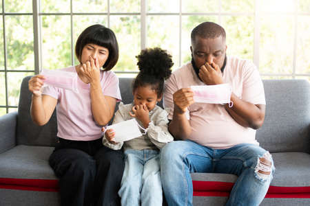 Mixed Race family sitting on the sofa and wearing medical face masks for against  world pandemic and stay quarantined together at home social distancing new normal lifestyleの写真素材