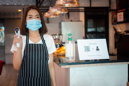 Asian waitress woman wearing face masks and holding an infrared forehead thermometer to check body temperature for virus symptoms of customers before entering the restaurant ( coffee shop ).の写真素材