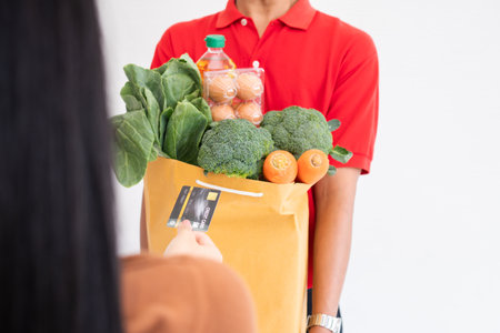 Asian delivery man from supermarket wearing a face mask and holding a bag of Fresh food, vegetables, and fruits for giving to customers at home. Concept of express grocery service and new  lifestyleの写真素材