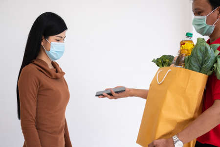 Asian delivery man holding holding a bag of fresh food for giving to customers and holding smartphone for Receive payments at home. Concept of express grocery service and new lifestyleの写真素材