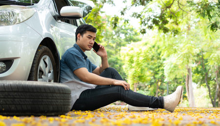 Asian man sitting beside car and using mobile phone calling for assistance after a car breakdown on street. Concept of vehicle engine problem or accident and emergency help from Professional mechanicの写真素材