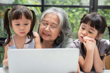 Asian Grandmother with her two grandchildren having fun and playing education games online with a computer notebook at home in the living room. Concept of online education and caring from parents.の写真素材