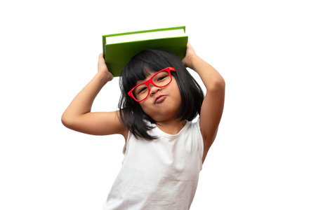 Funny and Happy Asian little preschool girl wearing red glasses holding a green book on the head, on...