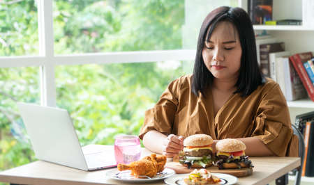Hungry overweight woman holding hamburger on a wooden plate, Fried Chicken and Pizza on table, During work from home, gain weight problem. Concept of binge eating disorder (BED).の写真素材