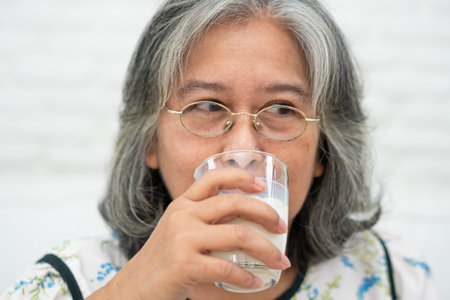 Asian Senior woman holding glass milk while relaxing on a sofa living room for retirement wellness. Elderly Woman drinking a glass of milk to maintain her wellbeing. Concept of wellness and healthy.の写真素材