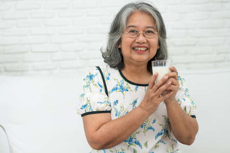 Asian Senior woman holding glass milk while relaxing on a sofa living room for retirement wellness. Elderly Woman drinking a glass of milk to maintain her wellbeing. Concept of wellness and healthy.の写真素材