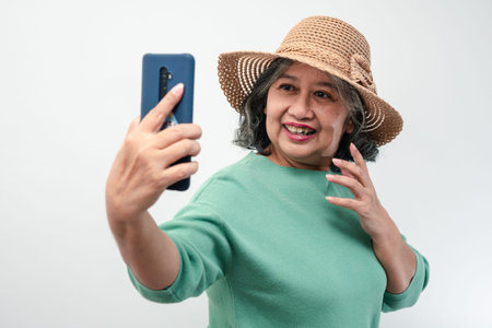 Portrait of happy Asian senior woman wearing sunglasses, a hat, and holding smartphone for a selfie,...