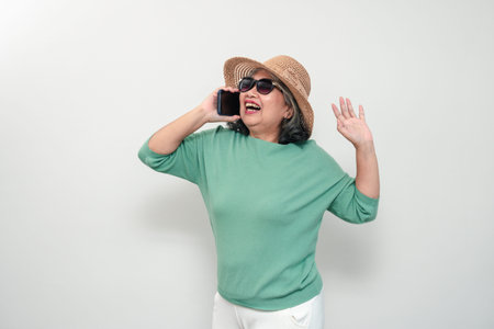 Portrait of happy asian senior woman wearing sunglasses, a hat, and holding smartphone standing...