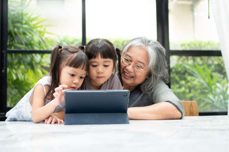 Asian Grandmother with her two grandchildren having fun and playing education games online with a digital tablet at home in the living room. Concept of online education and caring from parents.の写真素材