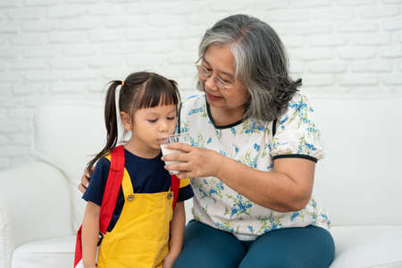 Happy elderly Asian Grandma sits beside her granddaughter and feeds fresh milk from glass for breakfast at home. Concept of a happy family and takes care together, preschool health careの写真素材