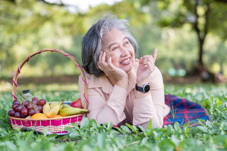 Happy Asian old senior woman and wear a health watch and lying on the picnic mat in park and basket of fruit besides. Concept of happy elderly woman after retirement and good healthの写真素材