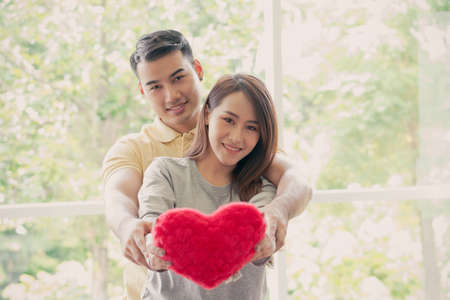 Happy couple sitting on the sofa and being a man embracing his girlfriend with love in the living room and smile and holding a red heart. Concept of romantic on valentine day. Proposal and marriageの写真素材