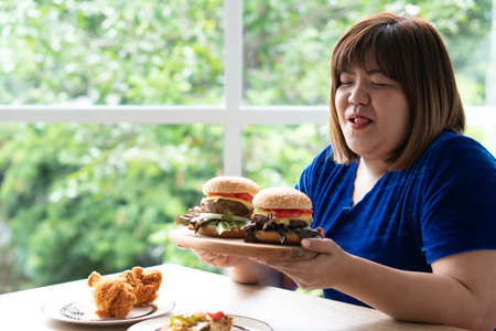 Hungry overweight woman holding hamburger on a wooden plate, Fried Chicken and Pizza on table, During work from home, gain weight problem. Concept of binge eating disorder (BED).の写真素材