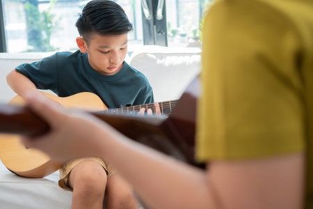 Asian boy playing guitar with father in the living room for teaching him son play guitar, feel...