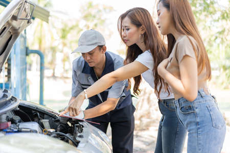 Asian male auto mechanic holds a clipboard and examines car engine breakdown problem with women customer and explain the root cause and estimate repair quotation, Car repair, and maintenance concept.の写真素材