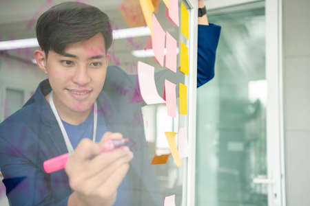 Young Creative businessman holding a marker and writing plan and share idea on glass wall with sticky note, Brainstorming and discussing and formulating, business strategies in tech startup office.の写真素材