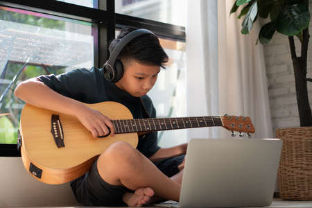Asian boy playing guitar and watching online course on laptop while practicing for learning music or musical instrument online at home. Boy students study online with video call teachers play guitar.の写真素材