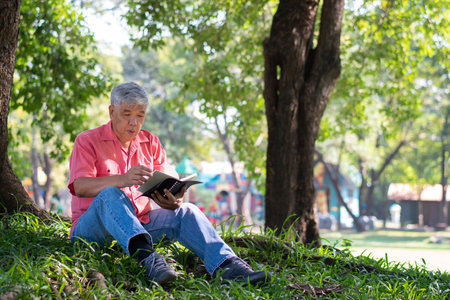 Happy Asian old senior man with gray hair reading book outside in park. Concept of happy elderly man after retirement and good healthの写真素材