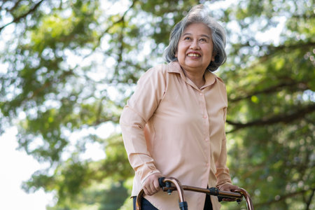 Happy old elderly Asian woman uses a walker for osteoarthritis rehabilitation physiotherapy in park.  Concept of happy retirement With care from a caregiver and Savings and senior health insuranceの写真素材