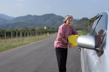 The car ran out of gas and stalled beside the road in suburbs and an elderly Asian woman used a gallon of spare gas to fuel the car. A woman prepares a gallon of spare gas to fuel before traveling.の写真素材