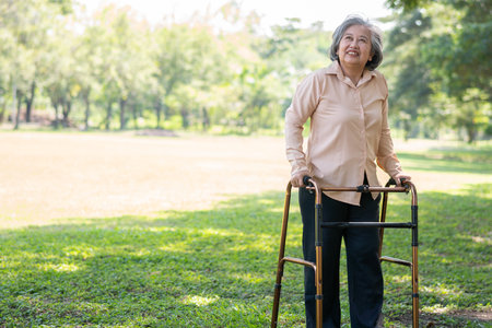 Happy old elderly Asian woman uses a walker for osteoarthritis rehabilitation physiotherapy in park.  Concept of happy retirement With care from a caregiver and Savings and senior health insuranceの写真素材