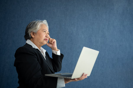 Portrait of intelligent senior businesswoman CEO manager holding in hands laptop creating a presentation on isolated blue background. Old Woman thinking after getting financial statements reports.の写真素材