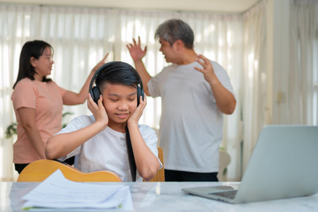 Asian boy kid wearing headphones and play loud music. so as not to hear quarrel while parents having fight or quarrel conflict at home. Unhappy problem in family, Domestic problems in the family.の写真素材