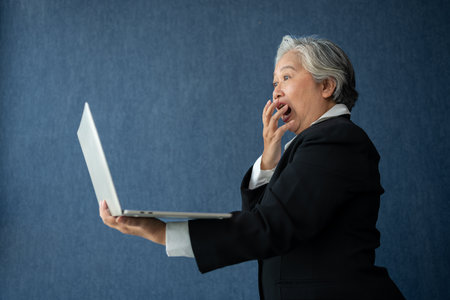 Portrait of intelligent senior businesswoman CEO manager holding laptop creating a presentation on isolated blue background. Old Woman is happy and smile after getting financial statements reportsの写真素材