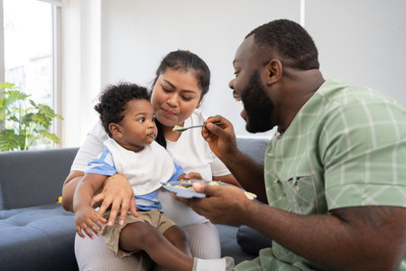 Asian mother feeding her 9 months old her cute little baby and African American helping for holding food plate At Home. Photo series of family, kids and happy people concept. Parents feed kids.の写真素材