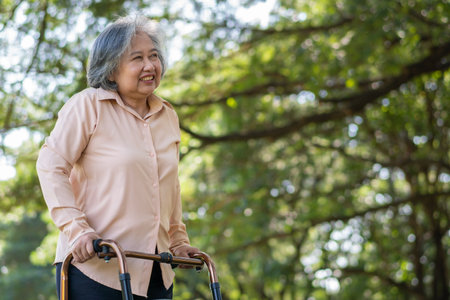 Happy old elderly Asian woman uses a walker for osteoarthritis rehabilitation physiotherapy in park.  Concept of happy retirement With care from a caregiver and Savings and senior health insuranceの写真素材