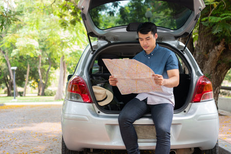 Asian man sitting back of car and holding map for find a place to travel. Happy road trip and vacation in summer in the countryside. Concept of journey on leisure and road trip vacationの写真素材