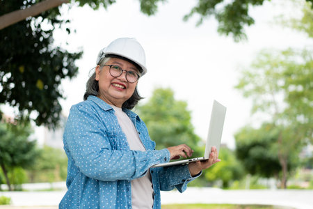 Happy elderly Asian professional engineer wears a helmet and checks the blueprint on a tablet on the...