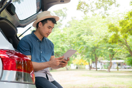 Asian man sitting back of car and holding tables check location and find place to travel. Happy road trip and vacation in summer in the countryside. Concept of journey on leisure, road trip vacationの写真素材