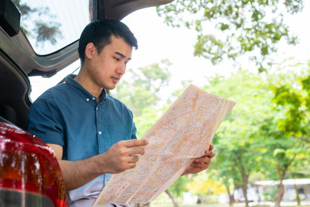 Asian man sitting back of car and holding map for find a place to travel. Happy road trip and vacation in summer in the countryside. Concept of journey on leisure and road trip vacationの写真素材
