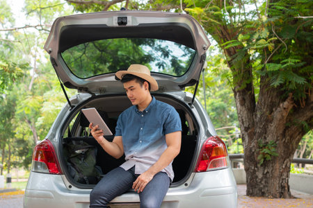 Asian man sitting back of car and holding tables check location and find place to travel. Happy road trip and vacation in summer in the countryside. Concept of journey on leisure, road trip vacationの写真素材