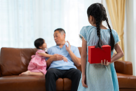 Little girl hiding present box behind back while making surprise elderly grandparents, Cheerful elderly man smiling and gesticulating while receives a surprise gift from granddaughter for celebrationの写真素材