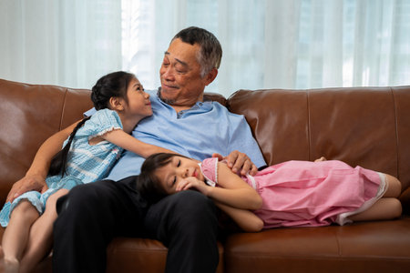 Happy Asian senior grandfather sits on a couch with his granddaughter and plays with his...
