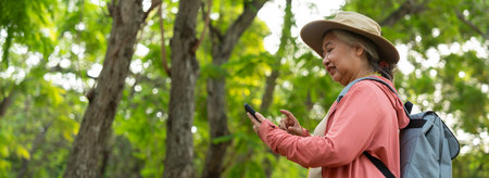 Portrait of Asian mature woman holding smartphone and backpack behind back, an Asia active senior woman enjoying nature in park. Standing on a trail in a forest outdoors. Enjoying active travel tripの写真素材