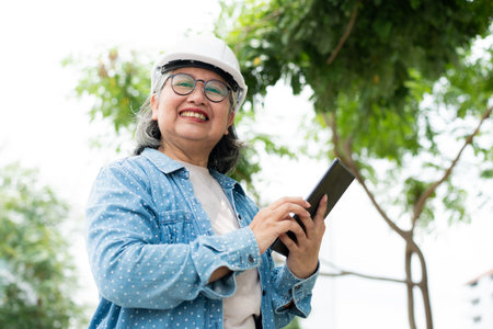 Happy elderly Asian professional engineer wears a helmet and checks the blueprint on a tablet on the construction site after the contractor and architect finish renovation the house and buildingの写真素材